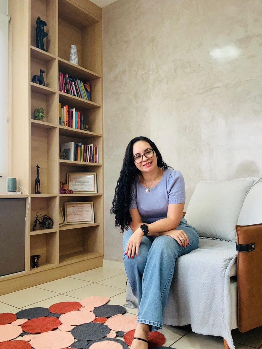 Laiane Andreoleti sentada em uma poltrona no consultório, ao lado de uma estante com livros e certificados.