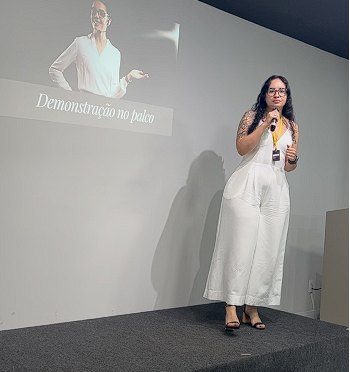 Laiane Andreoleti falando ao microfone em um palco, diante de uma projeção com o título "Demonstração no palco".